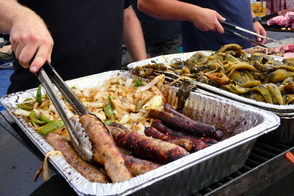 Two people hold tongs as they organize sausages and roasted chiles on trays