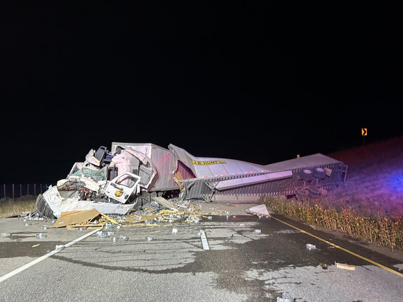 Two semi trucks crashed into each other, with major damage done to the front of one truck, including broken glass and a crushed cab, and major damage done to the body of the other truck, which is lying twisted on its side with debris scattered across the road.