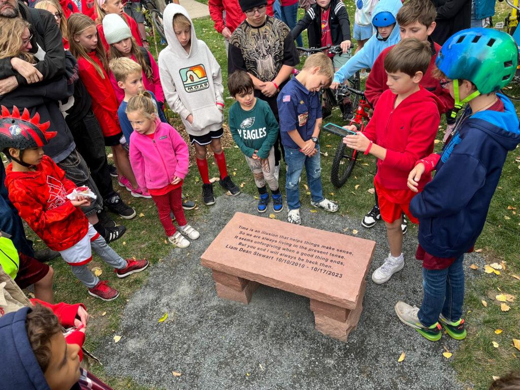 A crowd of children and families encircle a stone bench memorial for a young boy