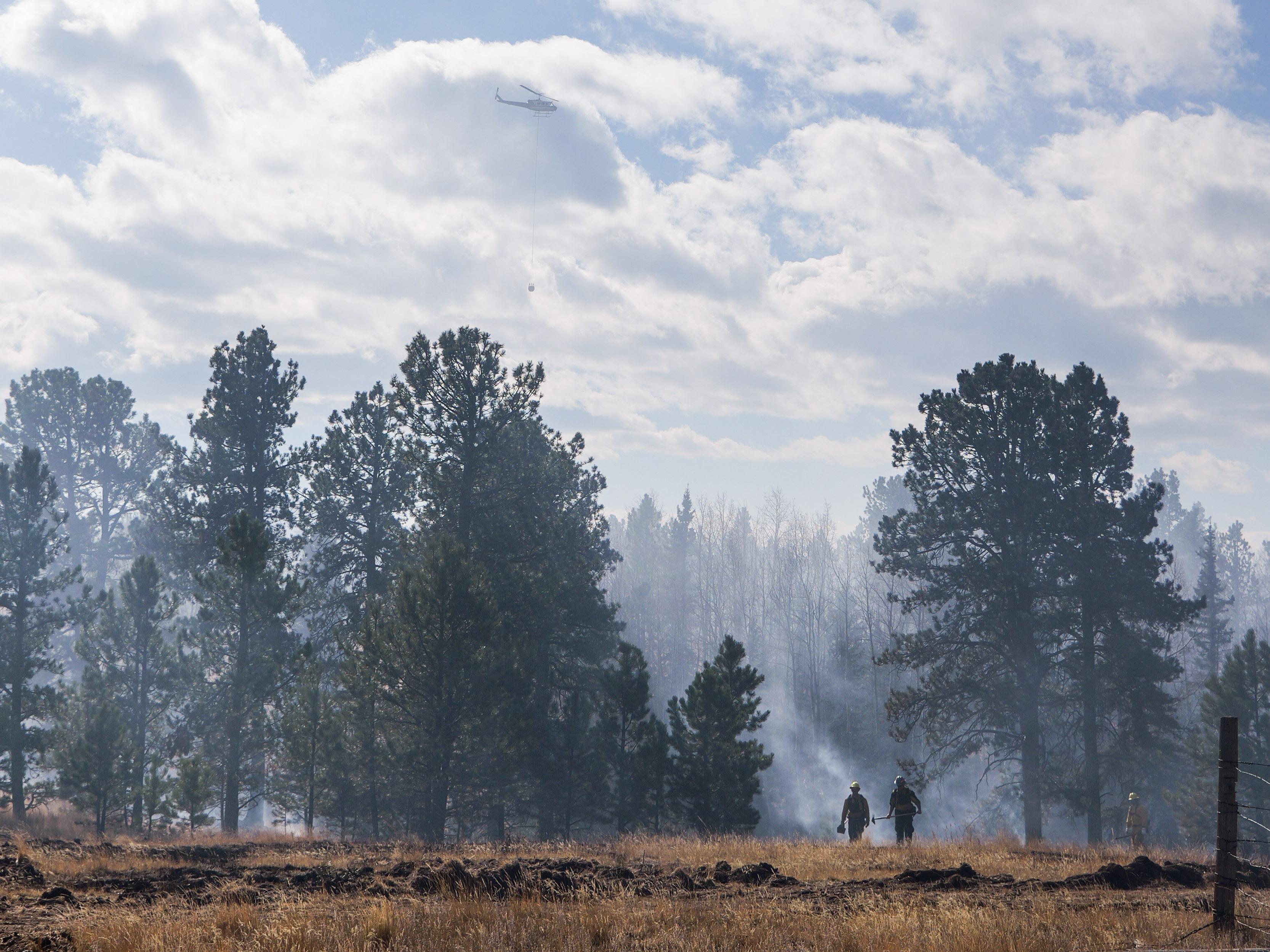 Firefighters walk through a smoke-filled forest with a tanker helicopter hovering above them