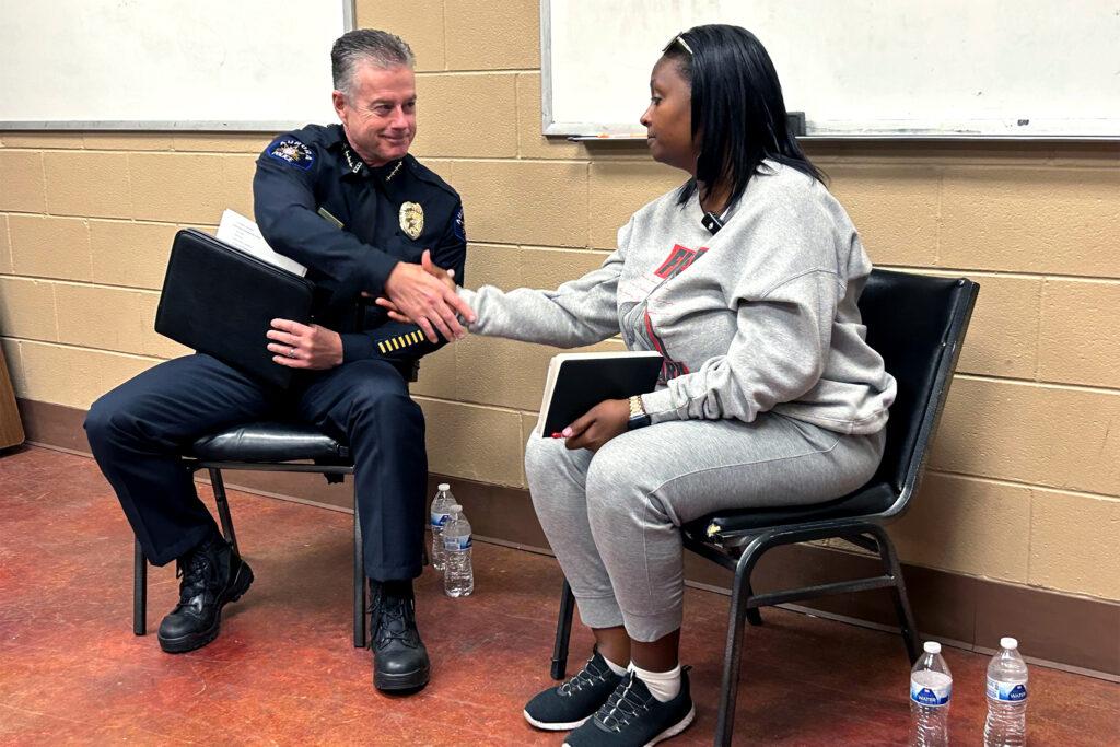 The police chief sitting on the right extends his right arm across his body to shake the hand of a woman seated next to him. The chief has a closed-mouth smile and the woman is not smiling.