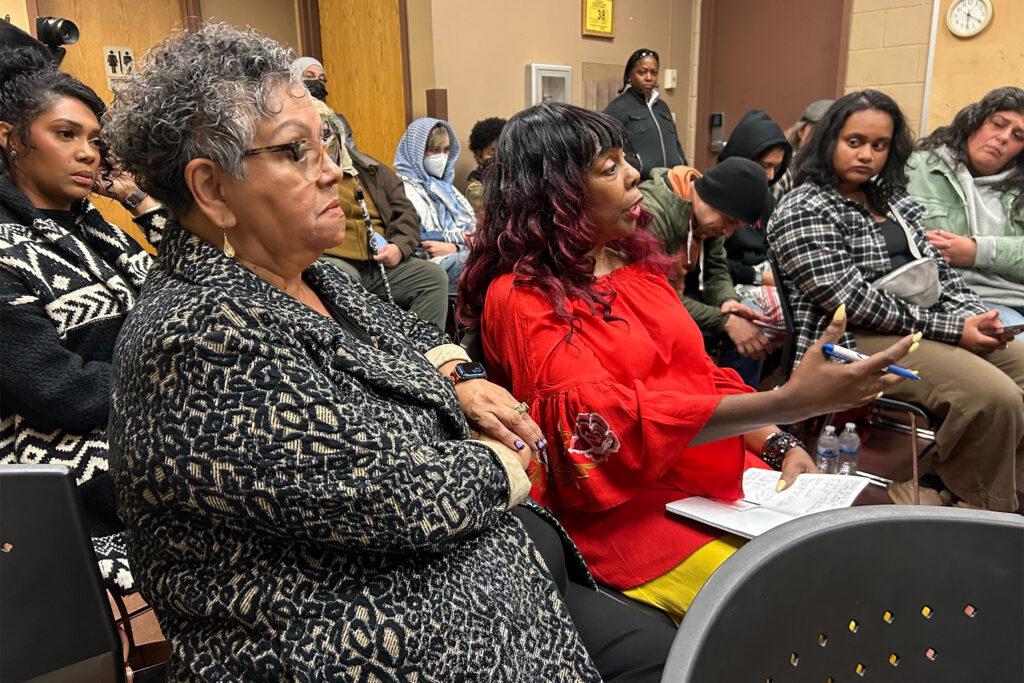 A side profile of two women sitting down in plastic chairs in the foreground as the one on the left gestures with her hand as her speaks. In the background are people listening.