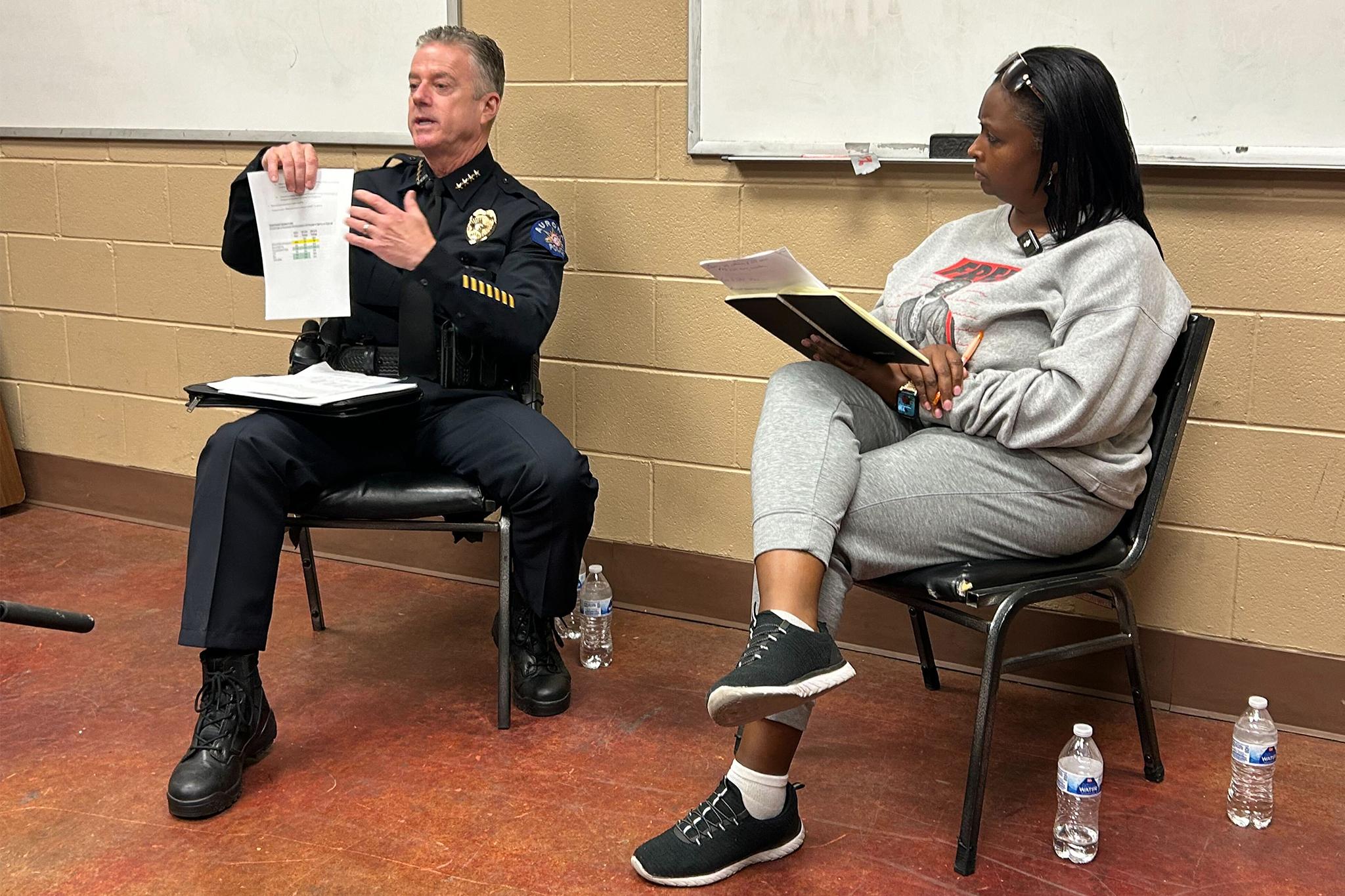 A police chief holds up a paper than shows statistics while he is sitting down. On the man's left is a woman who is also seated with her legs crossed. She is holding a notebook and pen and is looking at him with a quizzical look on her face.