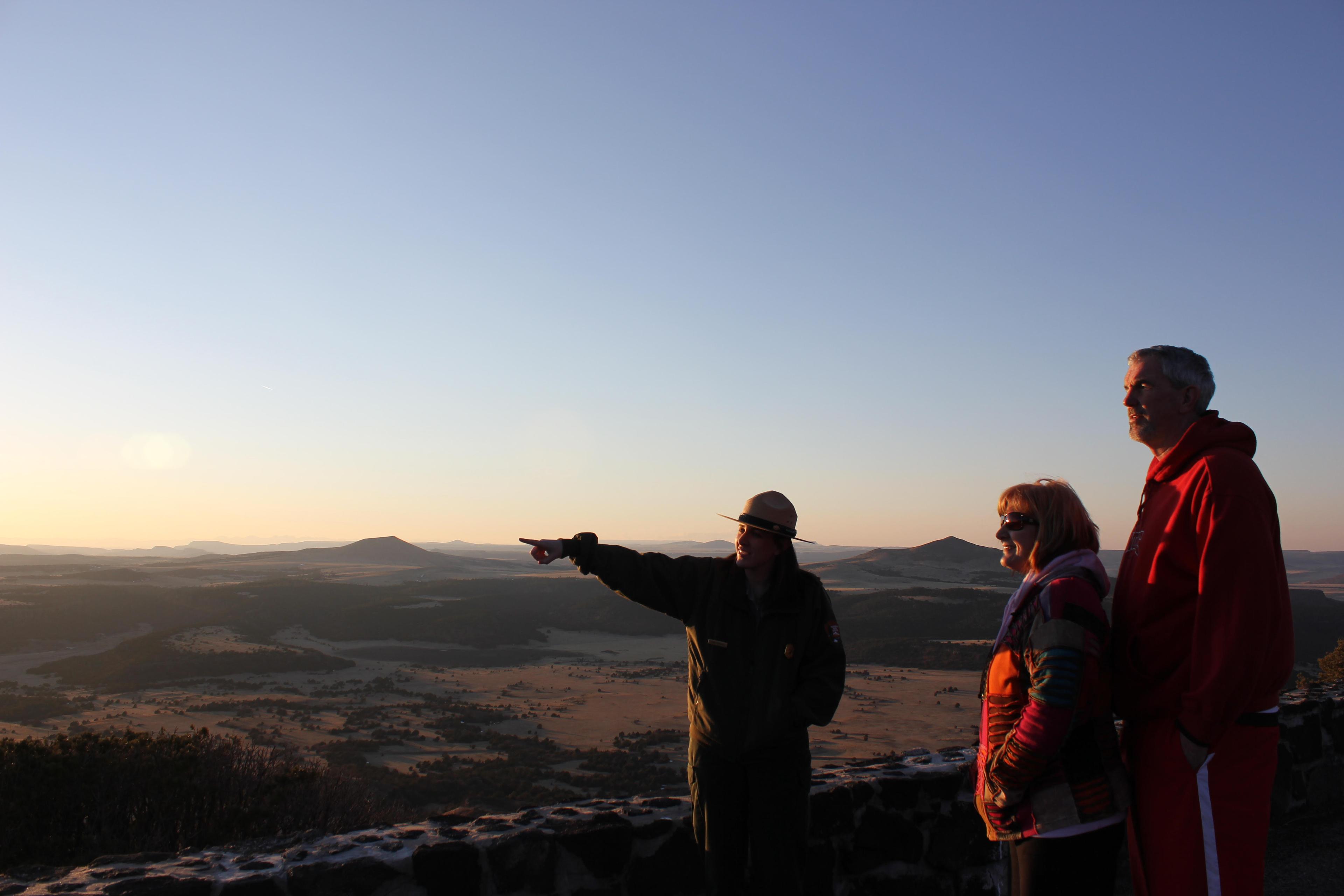 A park ranger points into the distance while talking to two people. There's a vast landscape in the background