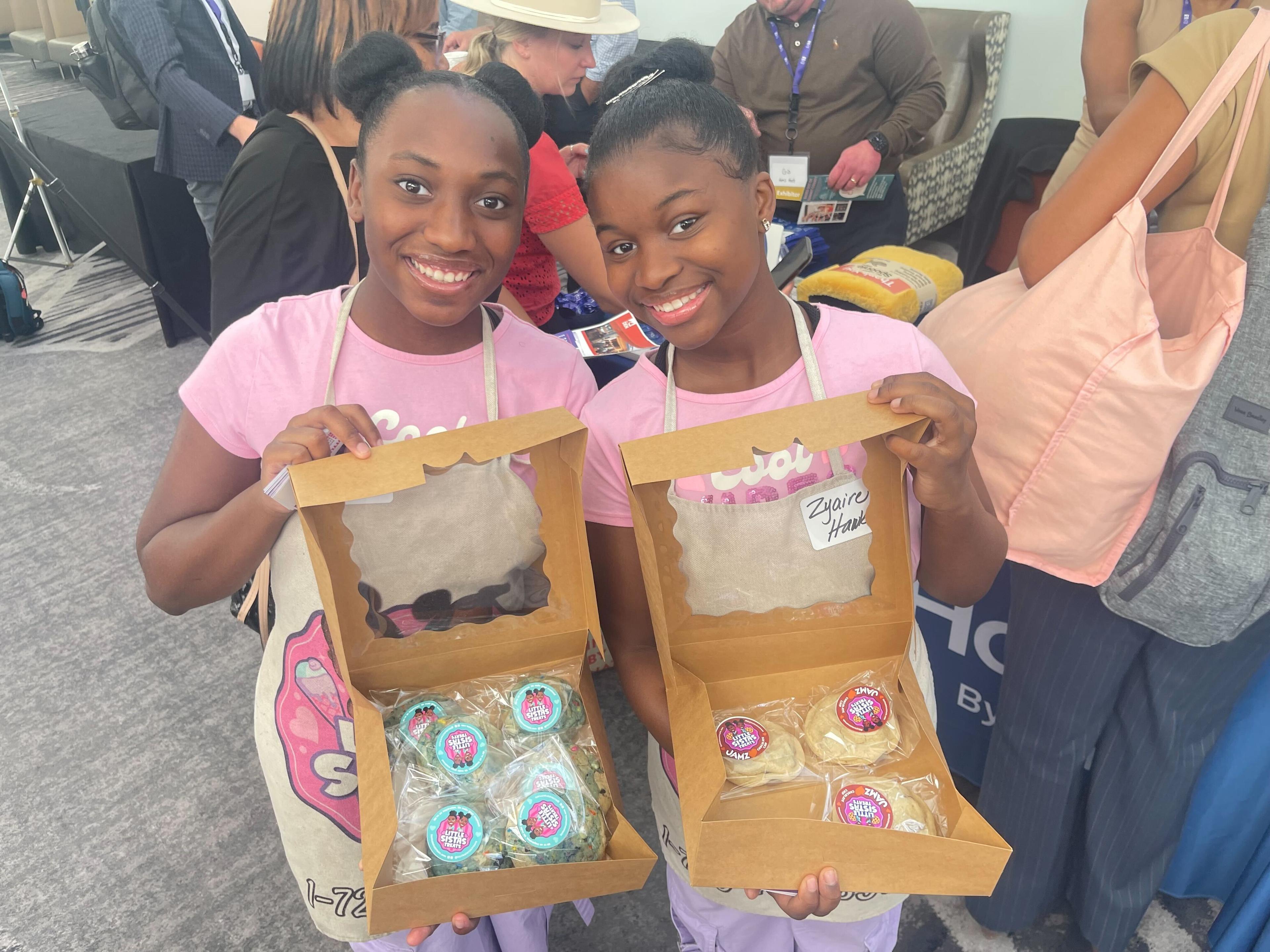 Twin sisters in matching pink shirts stand smiling with open boxes of their sweet treats.