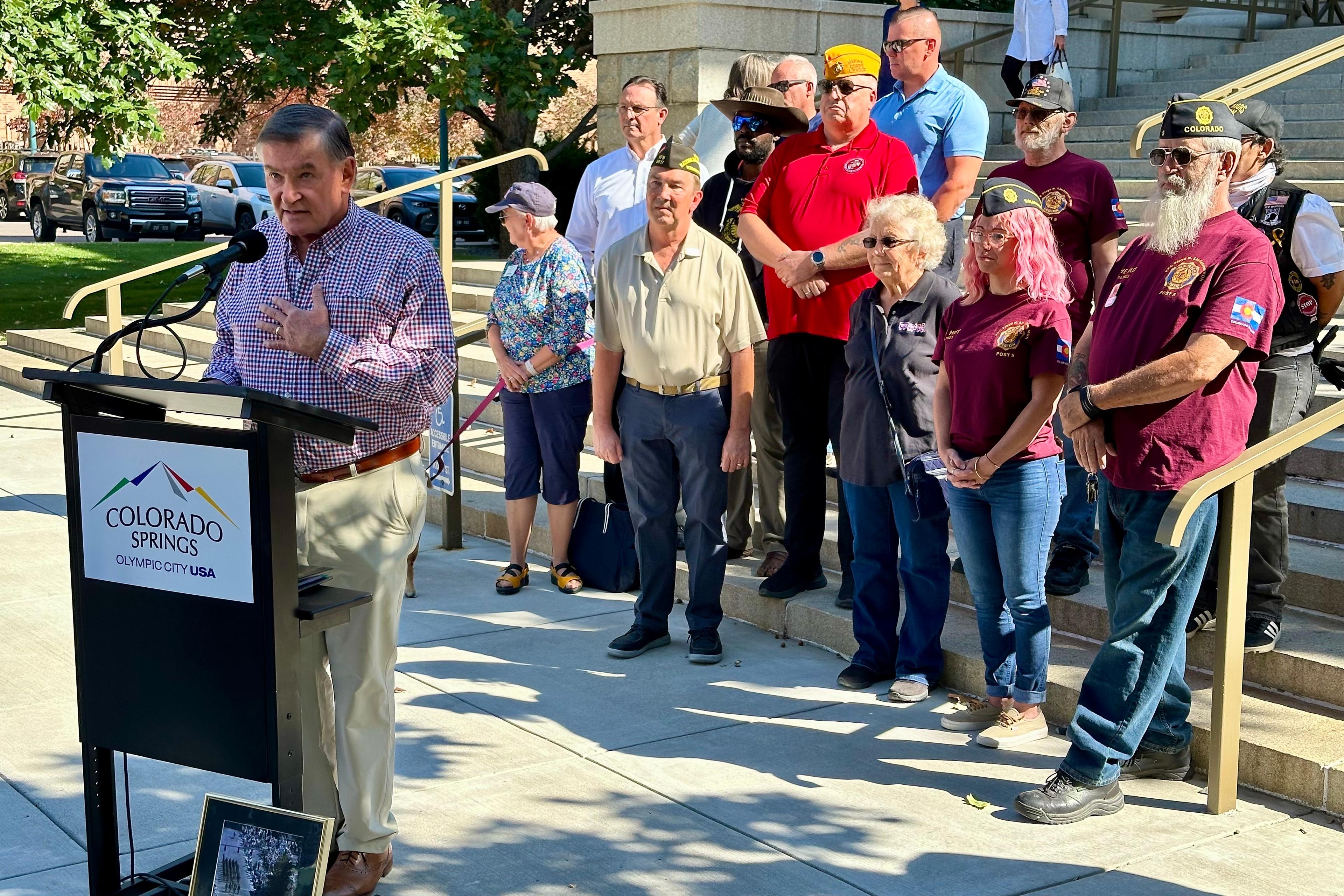 A man stands at a podium on the steps of Colorado Springs City Hall. A group of military veterans stand behind him.