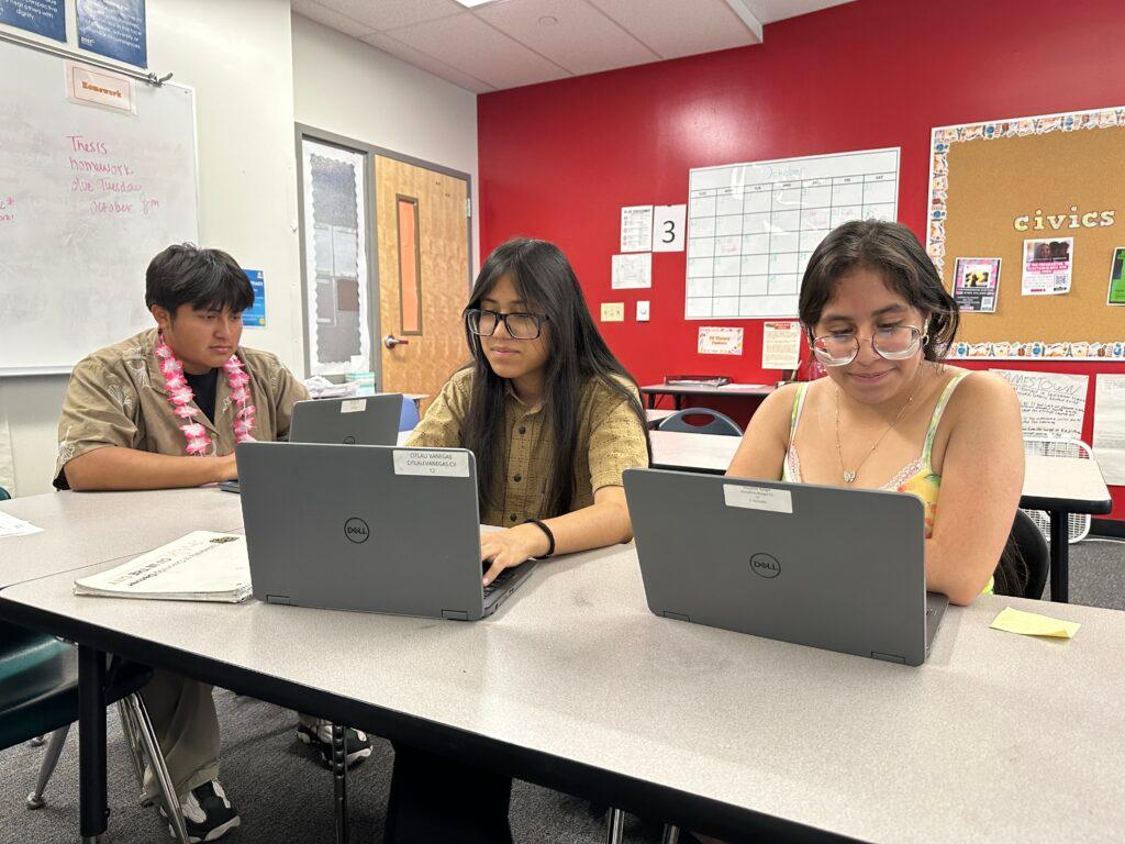 A high school boy wearing a lei and two high school girls sit at a white classroom table. All three students are looking at their Dell laptops.