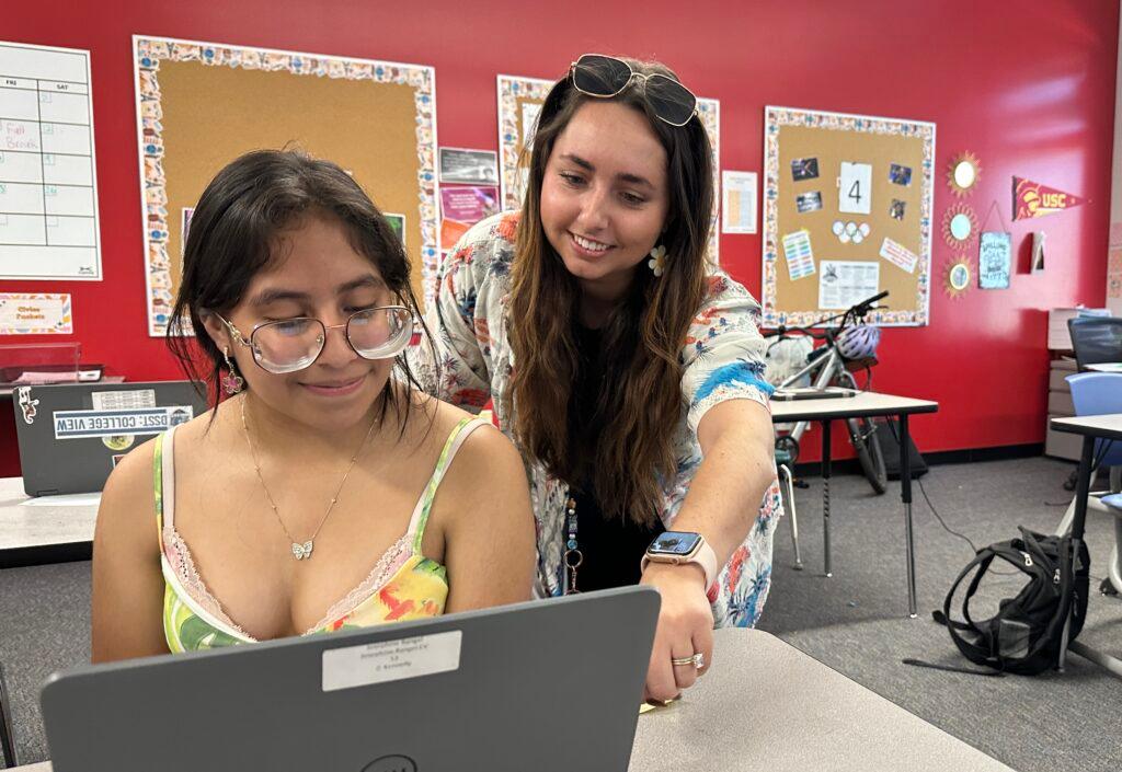 A teacher is smiling as she points to something on the laptop screen of a student who is seated.