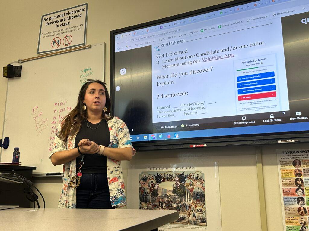 A teacher standing in front of a classroom in front of a big screen. The screen has an assignment on it that reads, &quot;Get informed. Learn about one candidate and/or one ballot measure using our VoteWise App. What did you discover? Explain.&quot;