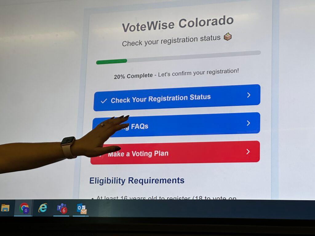 The picture shows the hand of a teacher pointing out something on a big screen that is displaying the VoteWise App. The app on the screen has two blue buttons and the teacher is pointing to the &quot;Check Your Registration Status.&quot;