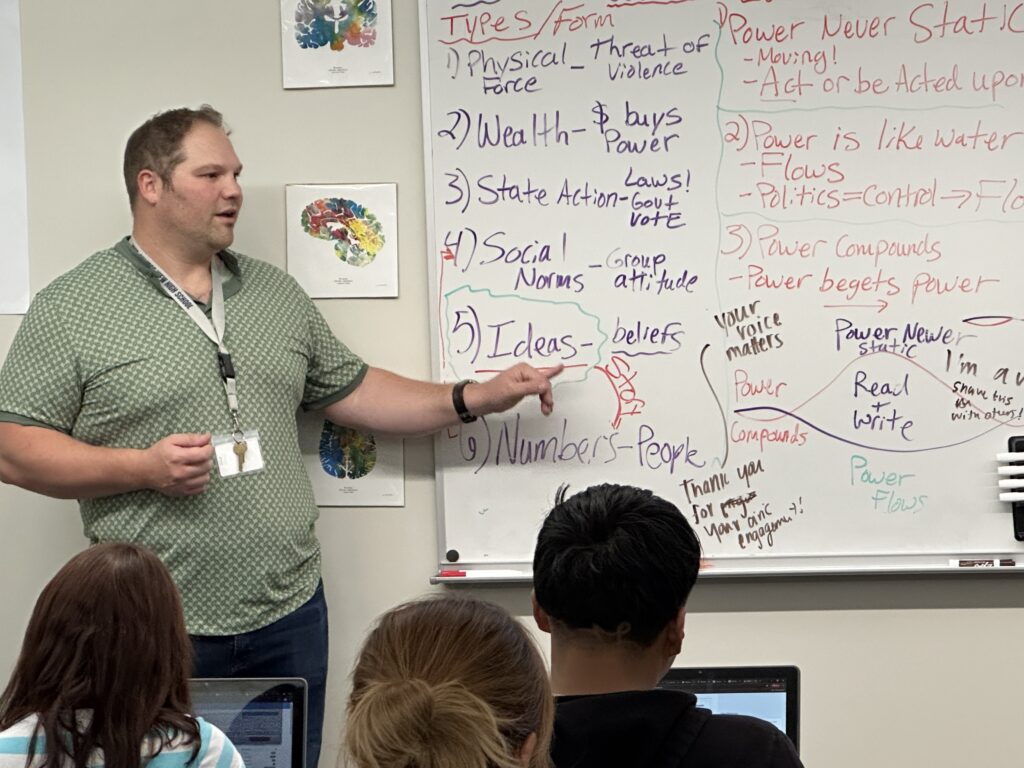 A male teacher points at a whiteboard in a classroom in front of students. On the white board, the words Physical Force, Wealth, State Action, Social Norms, Ideas and Numbers written in purple dry erase marker.