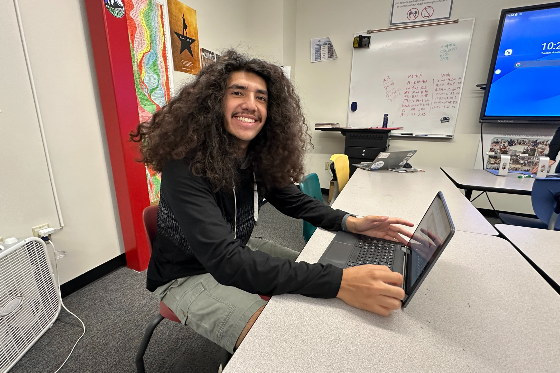 A high school student in the class sits at a white class table in front of his laptop and smiles at the camera. Behind him is a poster for "Hamilton," a white board full of class schedules and a sign that says "No personal electronic devices are allowed in class!"