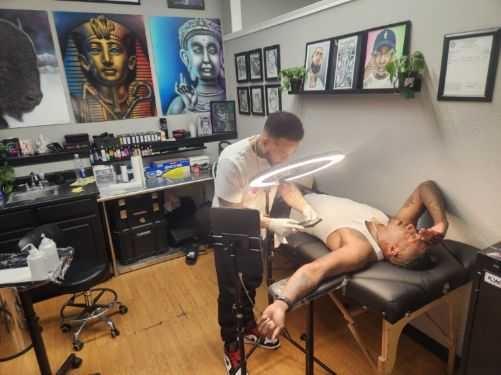 Photo shows a tattoo artist working on the arm of rapper Lecrae Moore who is lying down on a table in the tattoo shop in Aurora.