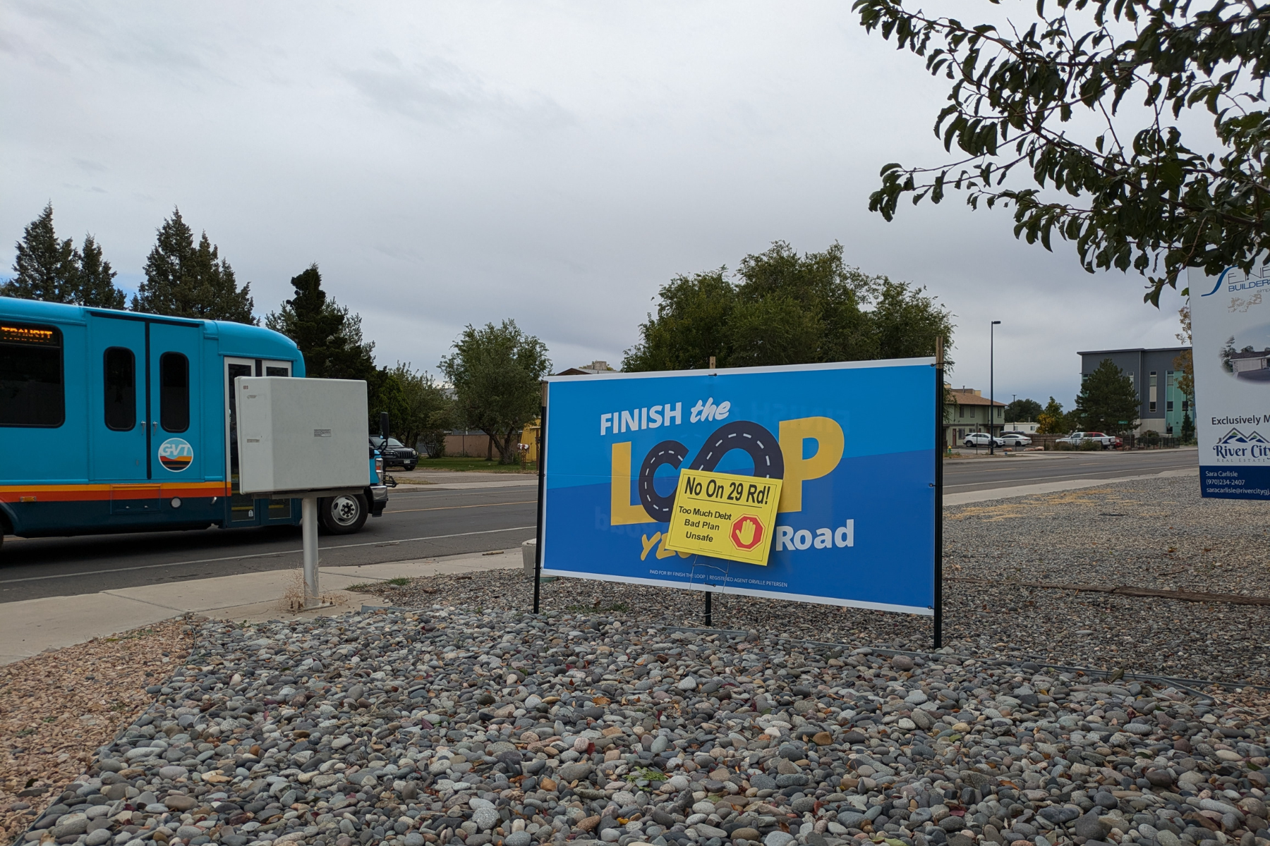 A turquoise Grand Valley Transit Bus, partially obscured by a mailbox, drives southbound on 29 Road past a pair of campaign signs for the 29 Road Interchange. A larger, blue sign is adorned with the slogan "Finish the Loop" while a smaller yellow sign sits in front of it reading "No On 29 Rd!"