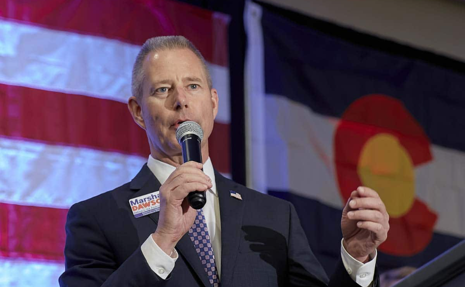 Marshall Dawson on stage with a microphone, standing in front of US and Colorado flags.