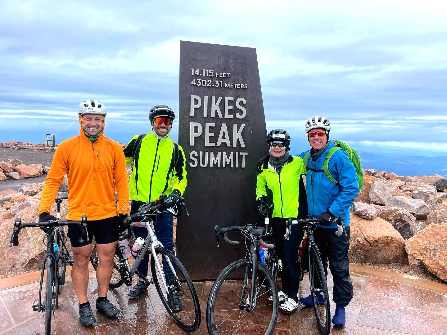 Four Cyclists with bikes stand next to the Pikes Peak summit sign