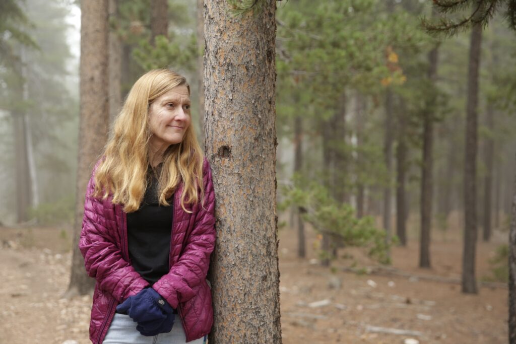 A woman in a purple puffy coat leans against a tree in a pine forest.