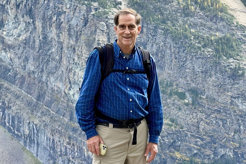 A man wearing a backpack stands in front of an alpine cliffside vista.