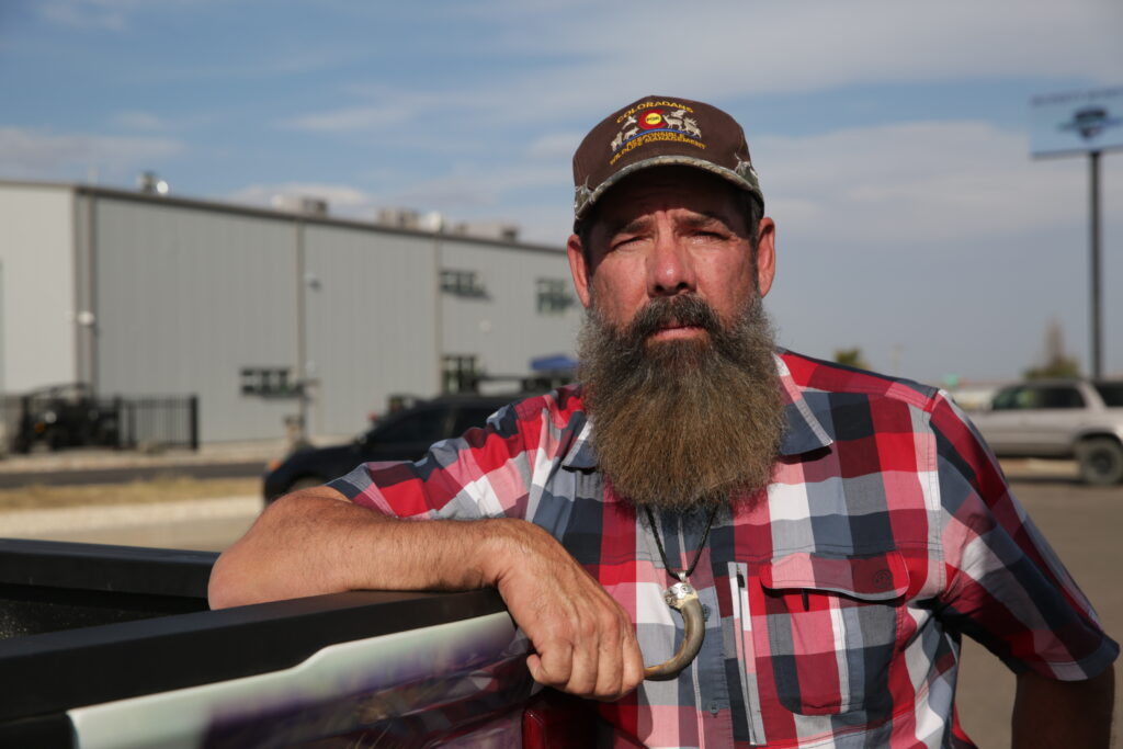 A man with a robust beard and ballcap leans on a truck tailgate. He wears a large bear claw necklace.