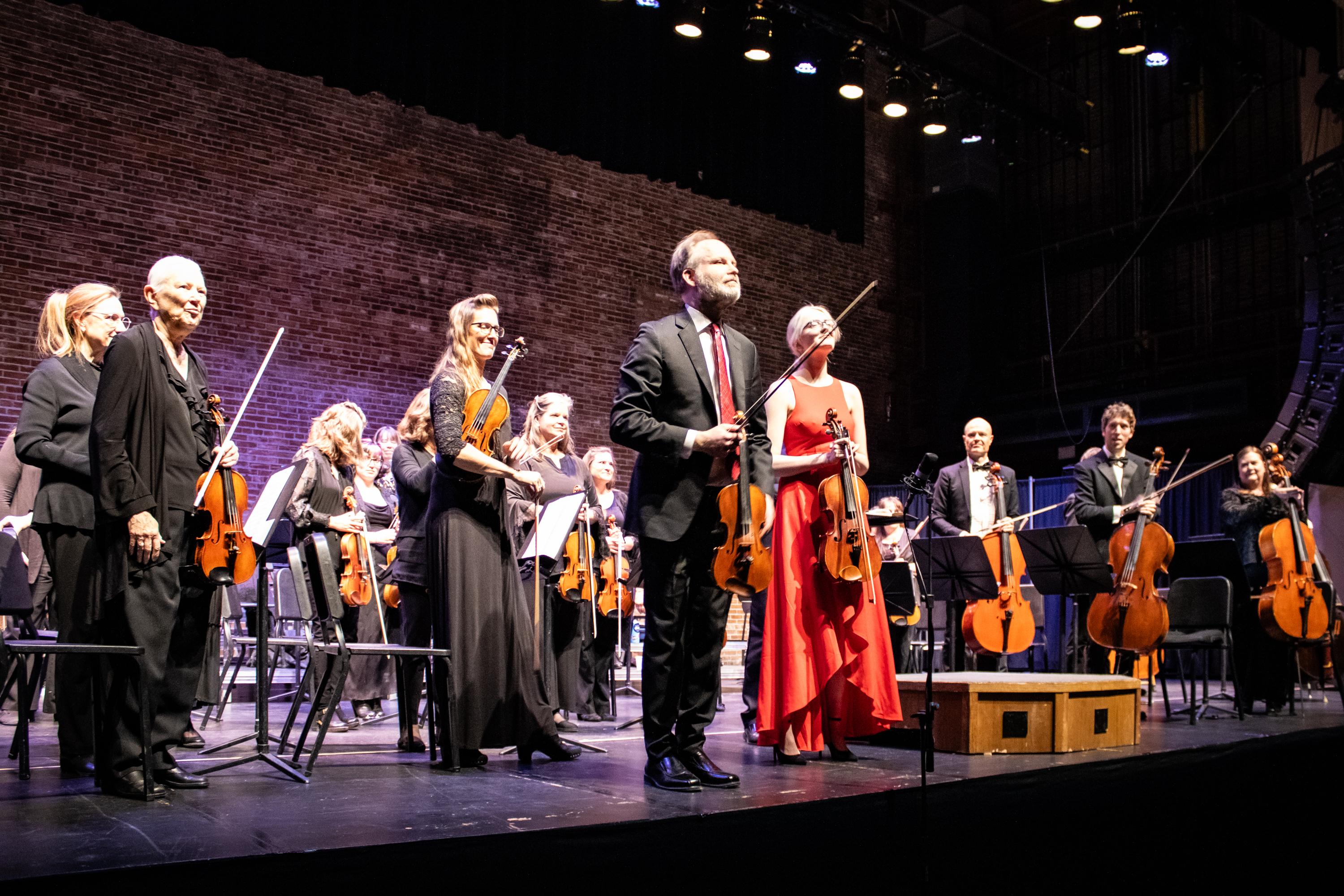 A man in a charcoal suit with a red tie holds a violin on stage while standing next to his daughter who is wearing a red dress.