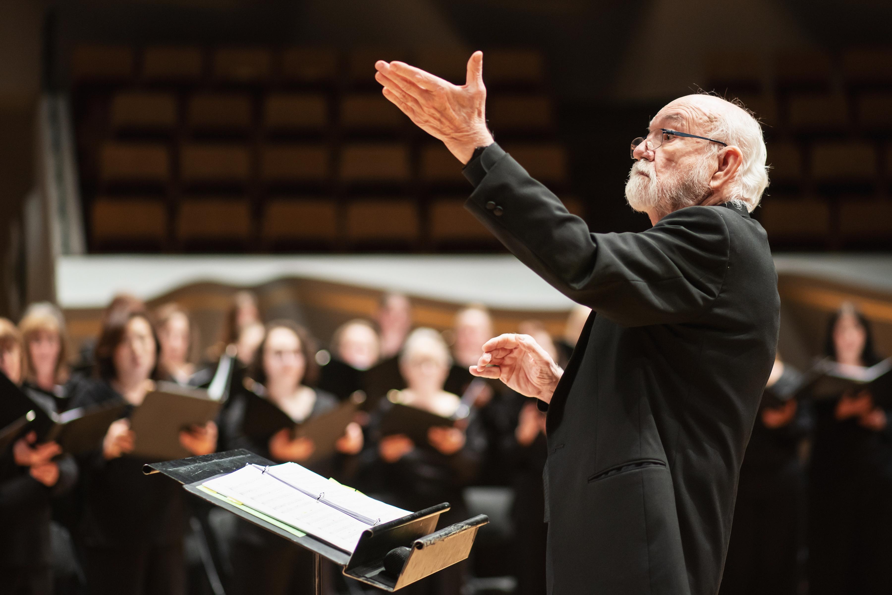 Conductor Duain Wolfe stands before a podium with arms raised, leading the Colorado Symphony Chorus.