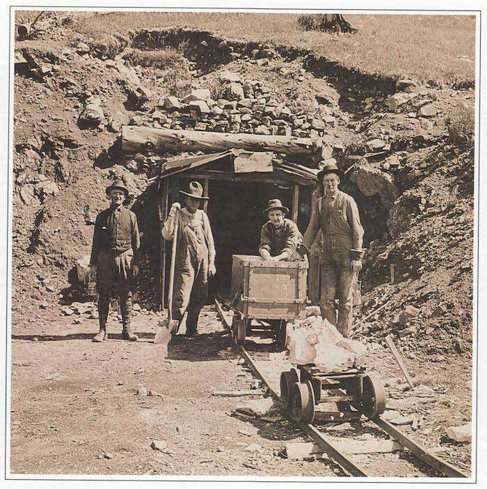 Miners stand near a track and an open entrance to a mine in an old sepia colored photo.