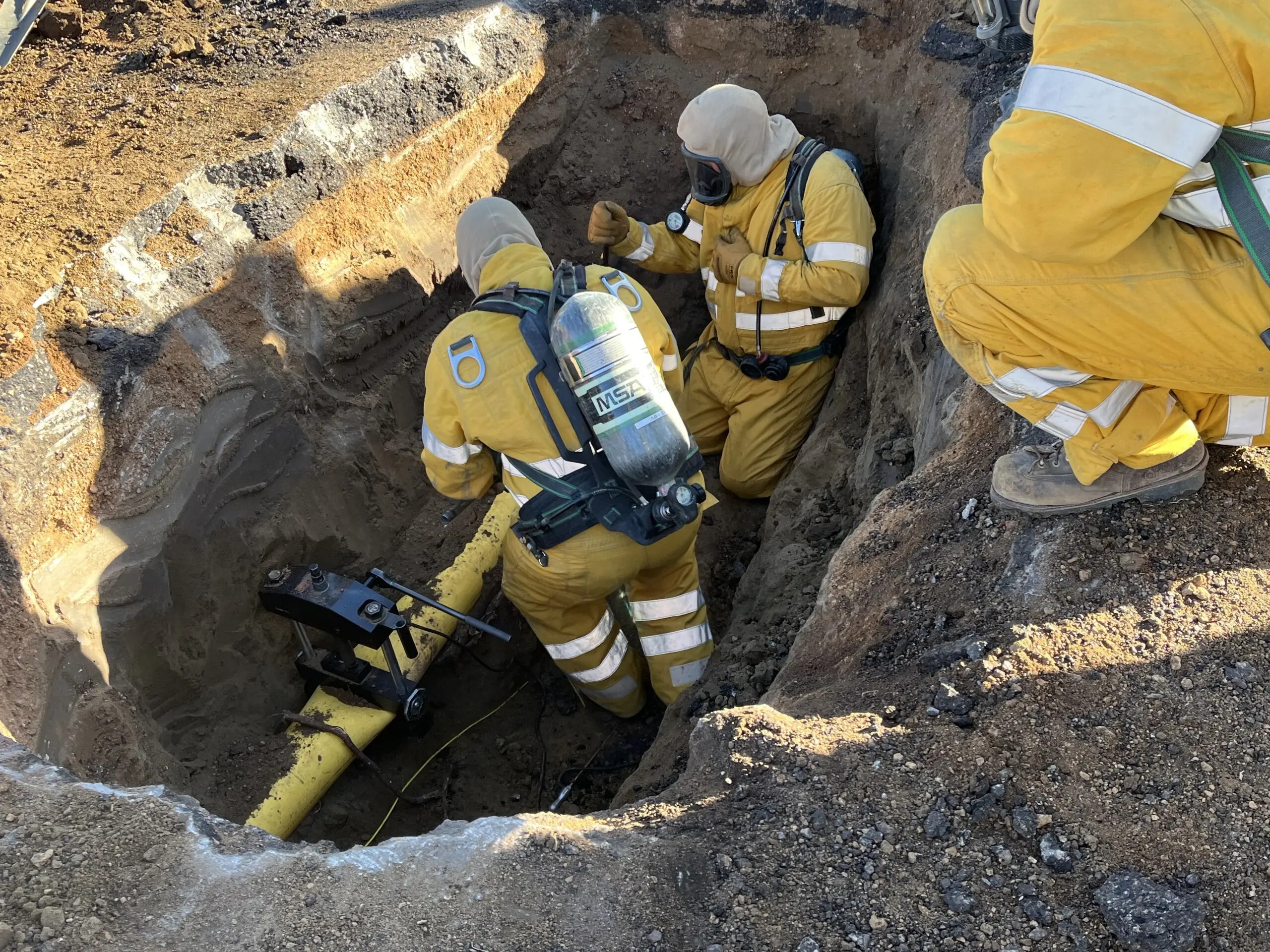 two workers wearing protective gear are in a trench working on a gas line while another watches nearby