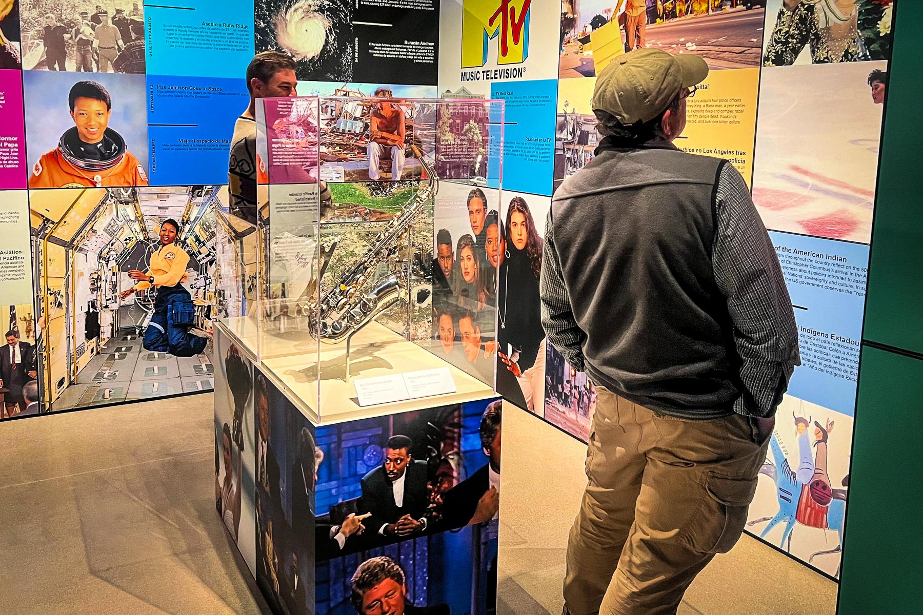 A silver and gold saxophone sits in a glass case in the center of a room wrapped with various images from the 1990s including astronauts, the MTV logo, and sitcom cast members. A man in a gray vest, hat, and tan cargo pants reads a portion of the exhibit near the saxophone.