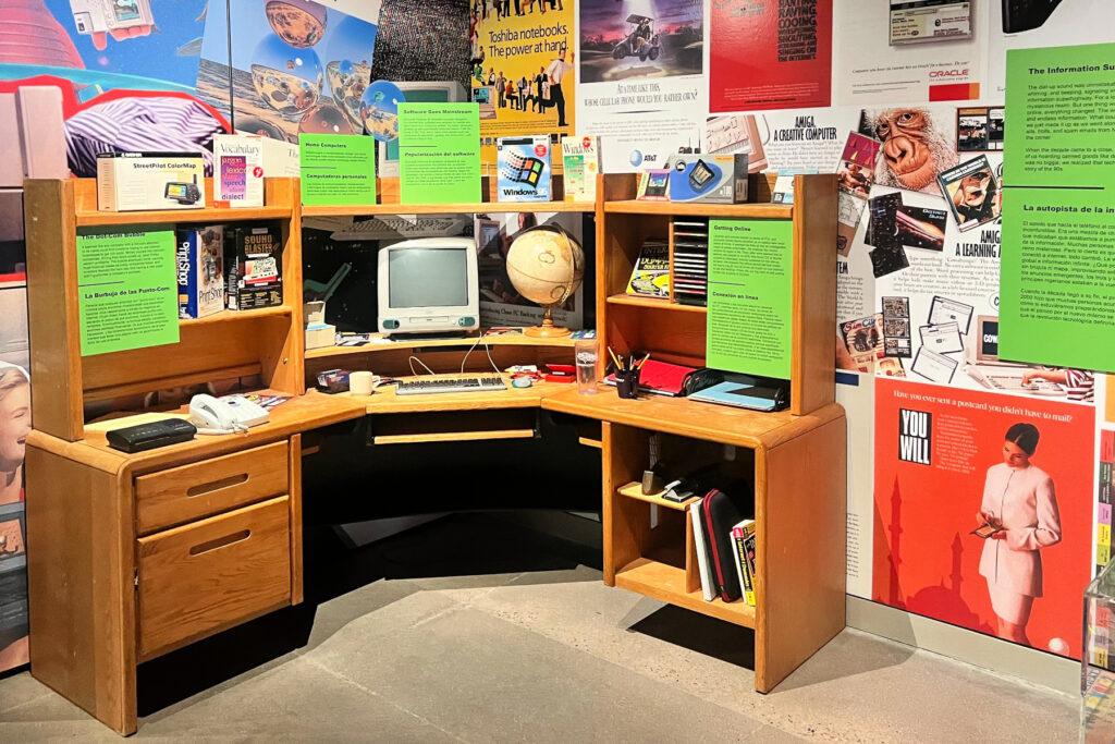 An oak wood desk displays a light blue and white MacBook desktop and other items from the era. The exhibit is modeled after a bedroom from the time.