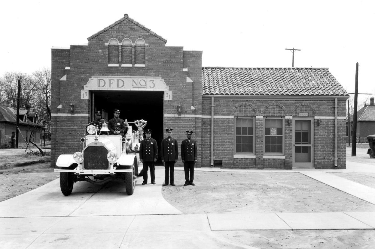 A black and white photo of 5 firefighters posed outside a brick fire station labeled D.F.D. No. 3. Two of the firefighters stand in an old fire vehicle facing the camera and three more stand adjacent to the vehicle with arms by their sides.