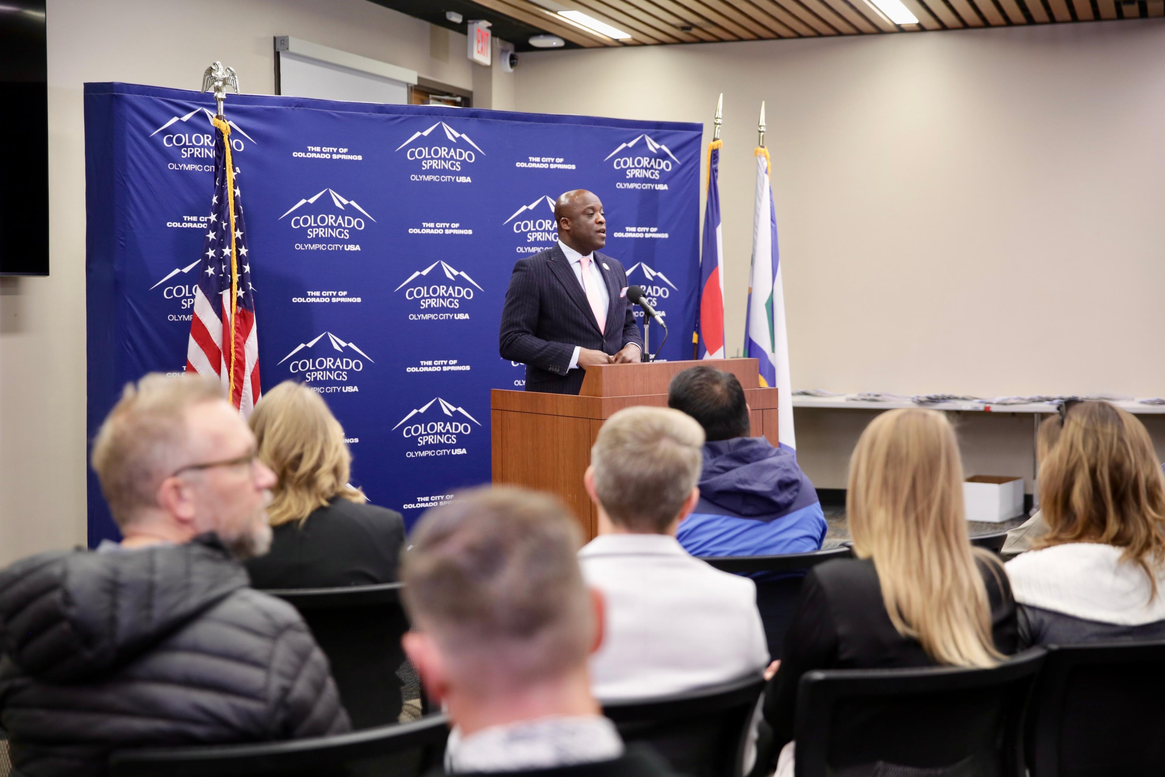 A man in a dark suit stands at a podium in front of a small group.