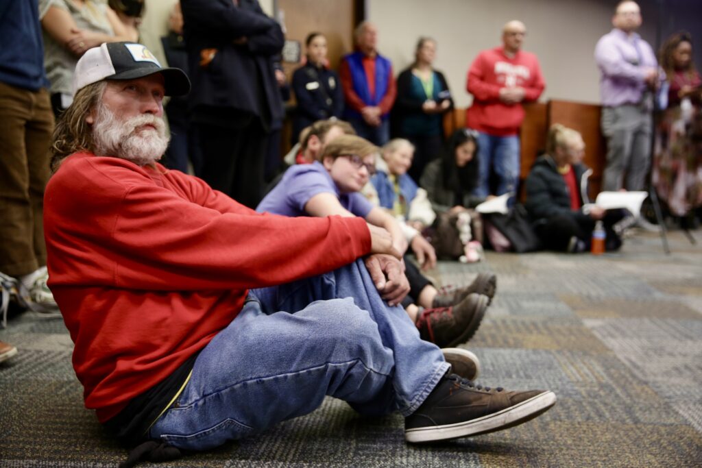 A man in a red sweater and baseball cap sits on a carpet amongst others doing the same.