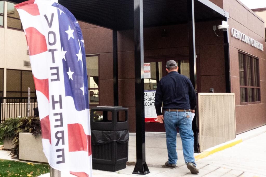 a person brings a ballot into a red colored entrance with a banner out side that say vote here