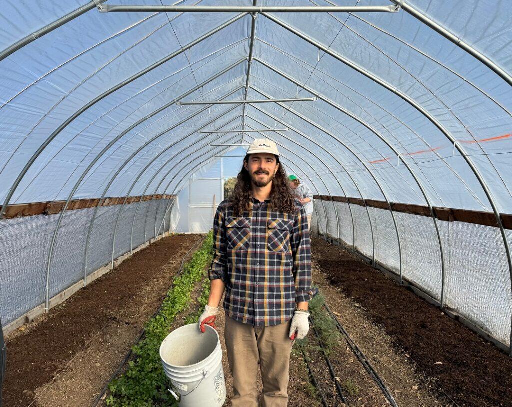 A man holds a bucket while standing inside a greenhouse