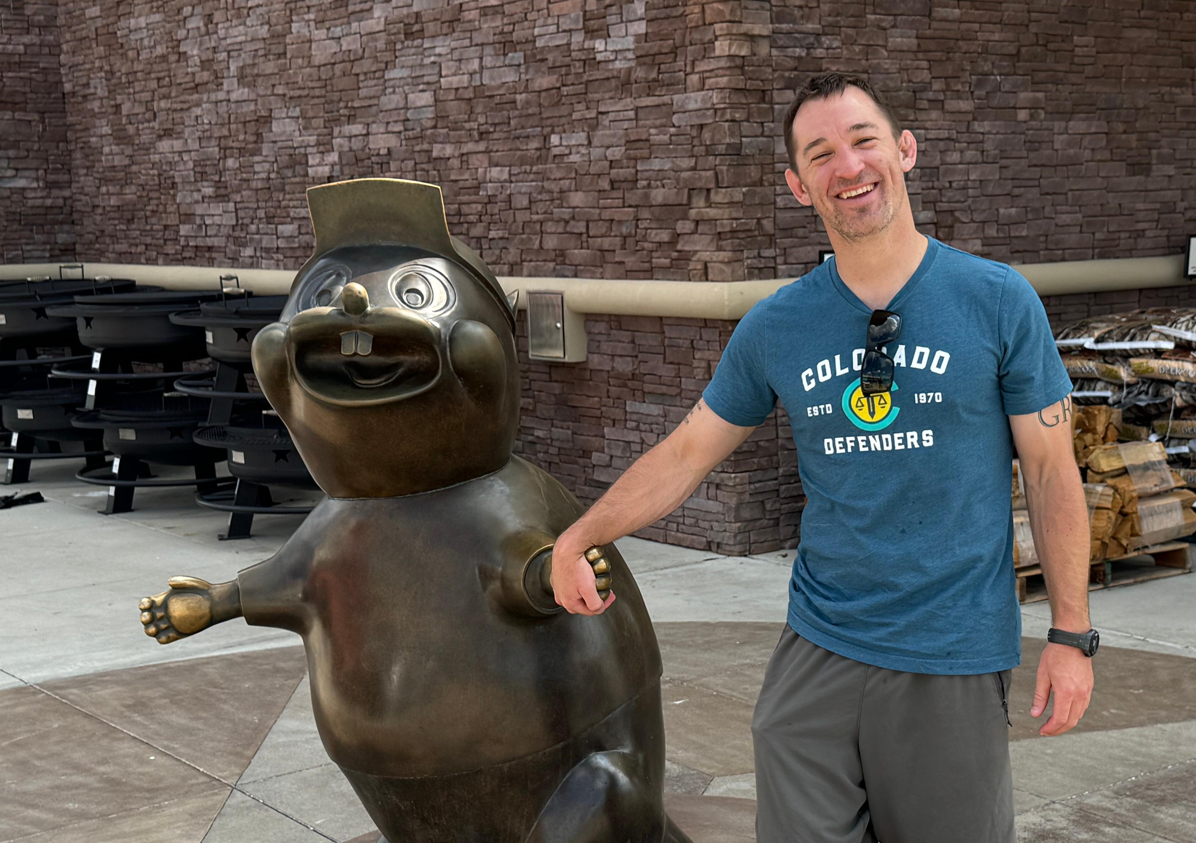 A man stands smiling as he holds the bronze hand of a statue of a beaver -- Bucky the Beaver, the convenience store Buc-ee’s mascot. The man is wearing a blue shirt that reads "Colorado Denfenders."