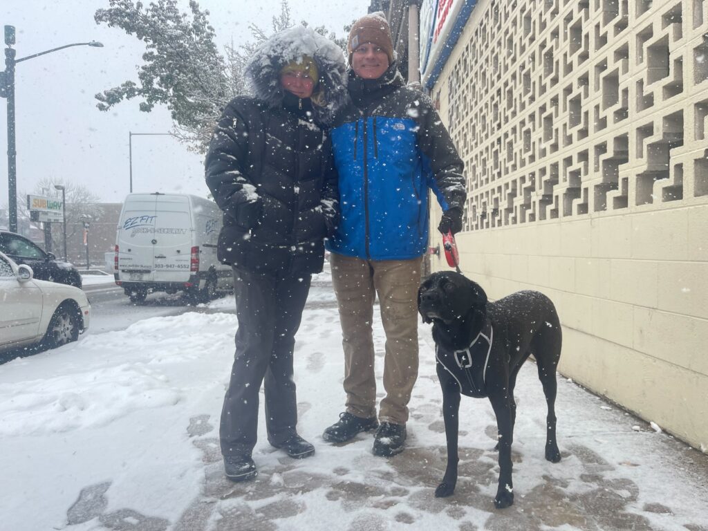 A couple bundles up to walk their old black dog in the snow.