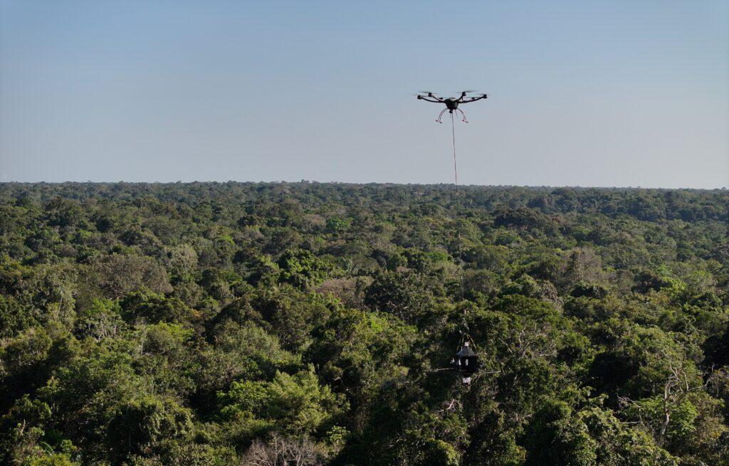 A drone flies over a rainforest canopy in Brazil carrying a piece of equipment suspended from a cable below.