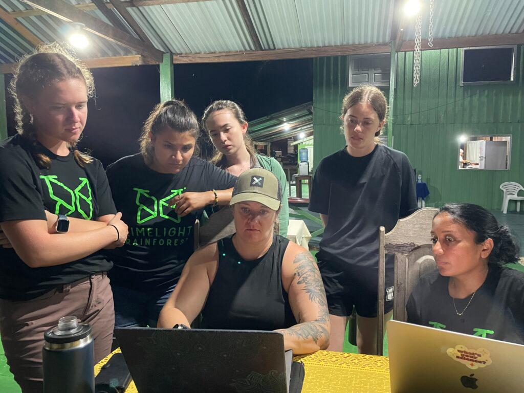 Six women gather around a laptop computer to examine DNA data.