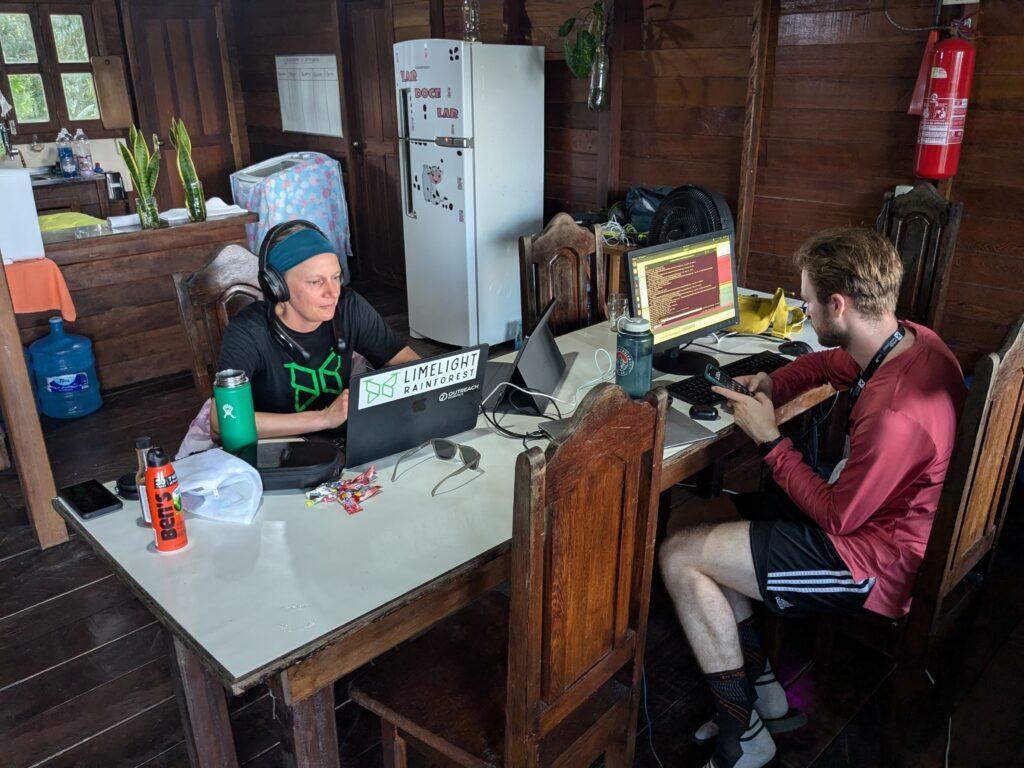 In a busy cabin with wood finishings a woman in a black shirt works on a laptop while a man in a red shirt on the opposite side of the table operates his calculator app on his phone while observing readings on a computer moniter.