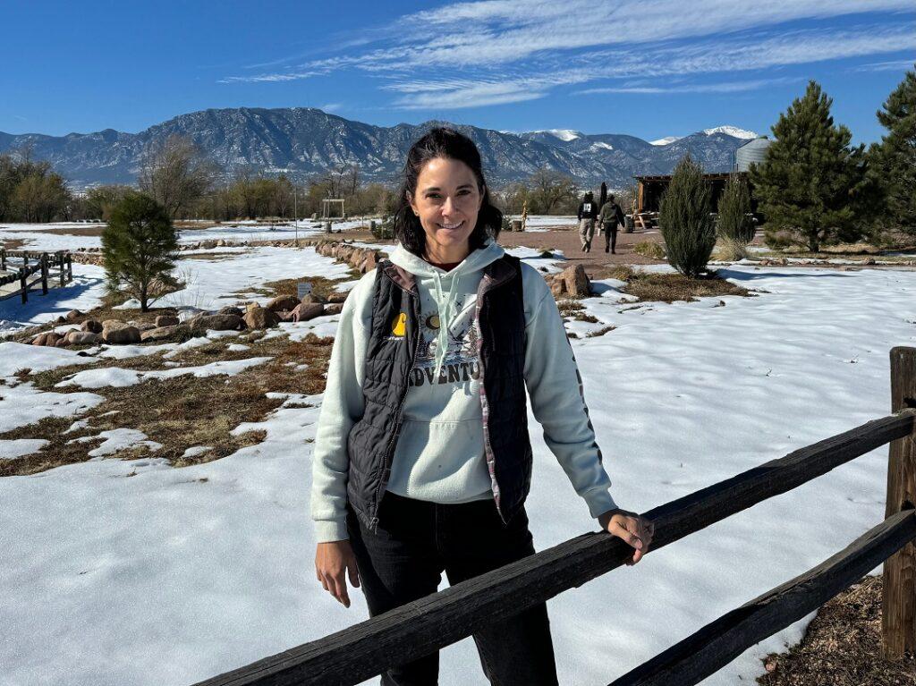 A woman stands next to a wooden fence with a snow-covered farm behind her.