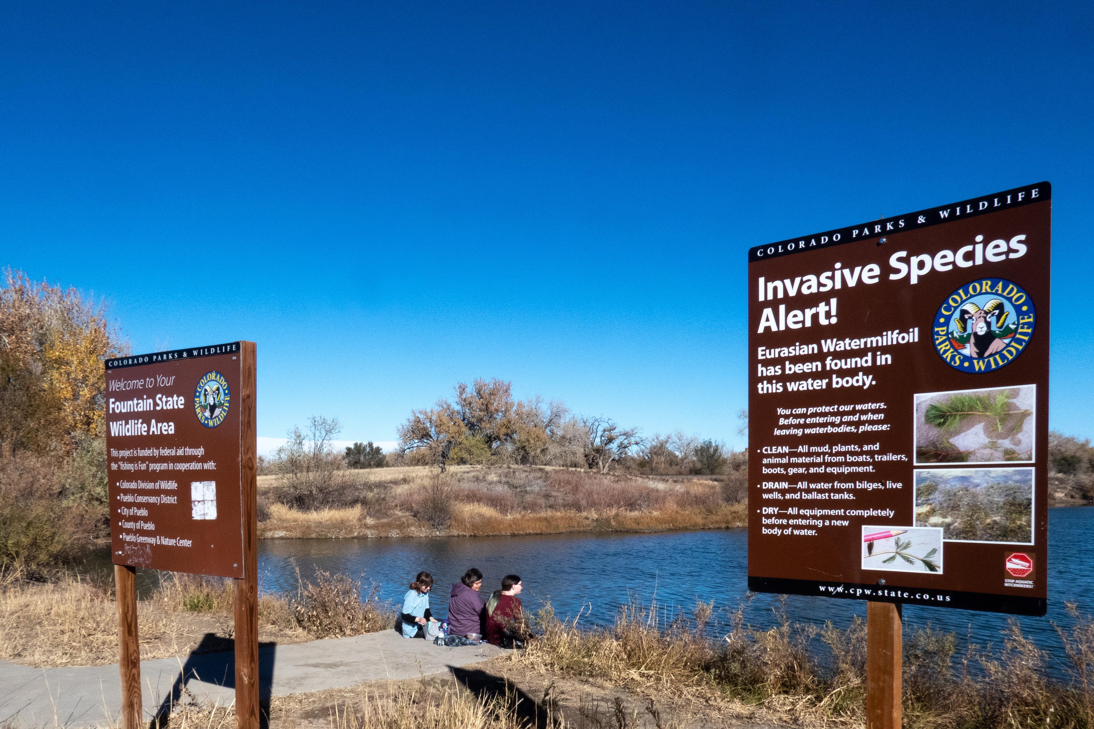 signs stand in front a small lake and three people sit nearby