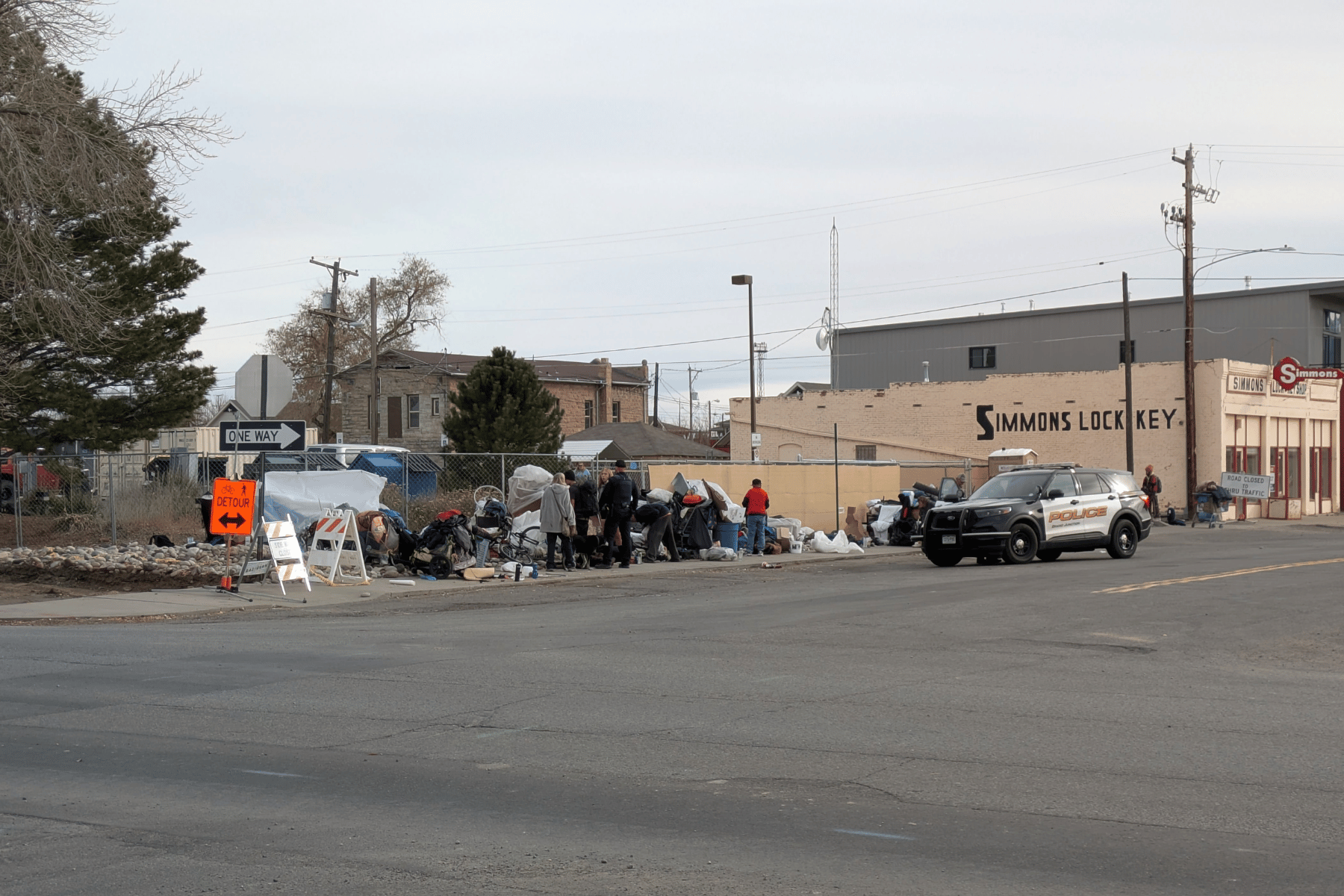 A Grand Junction Police Department car is parked in the street in front of a sidewalk that is crowded with people and their things. Grand Junction police officers speak with some residents on a closed sidewalk.