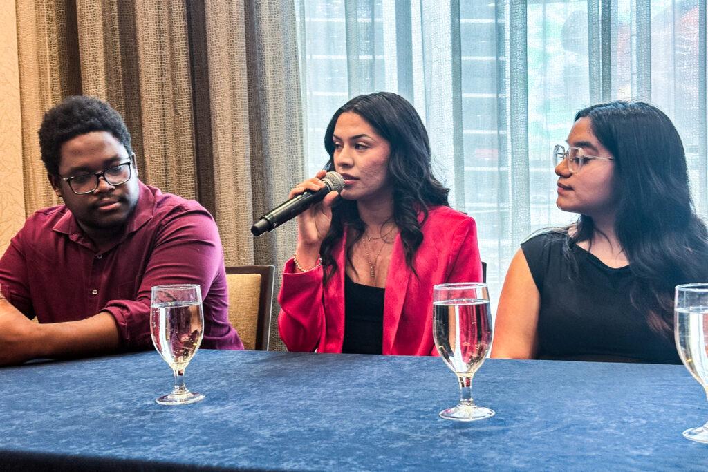 A student with a microphone is flanked by two other students in front of a table