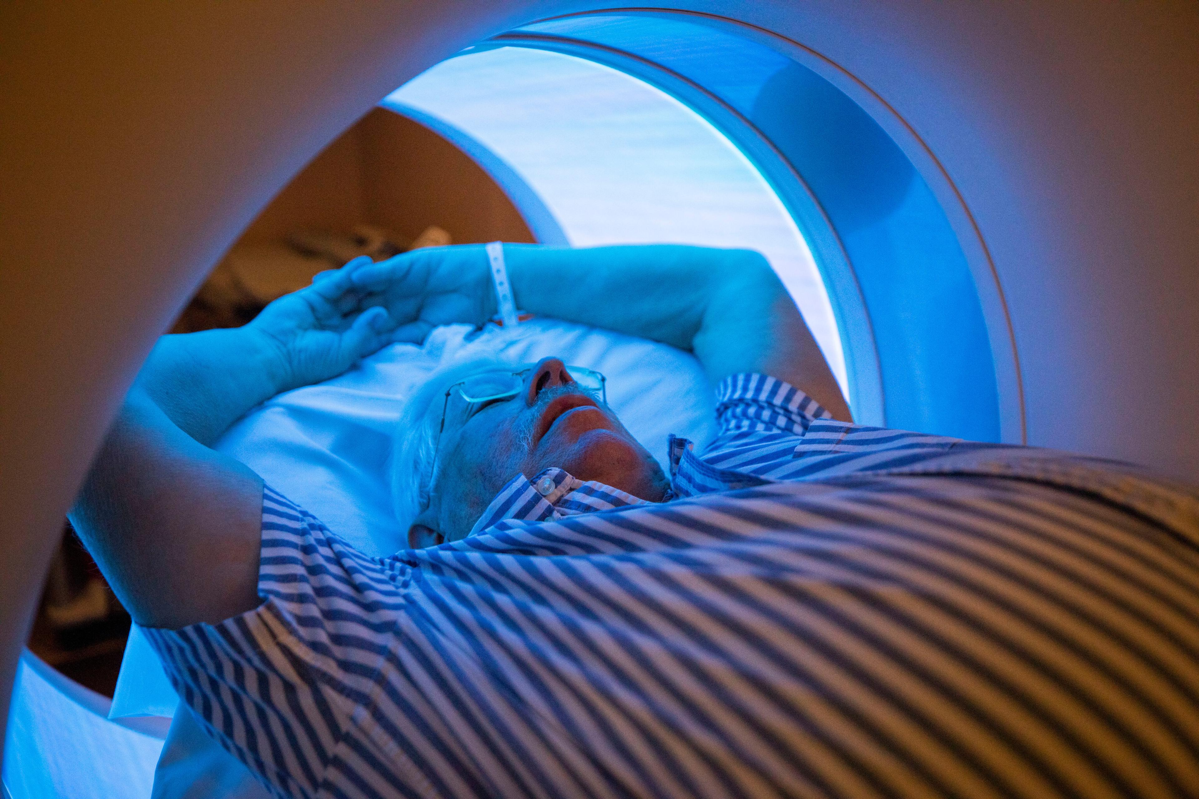 A close-up pf a man as he gets a CT scan. The light in the machine casts a blue hue on the man;s face and arms that are stretched above his head.