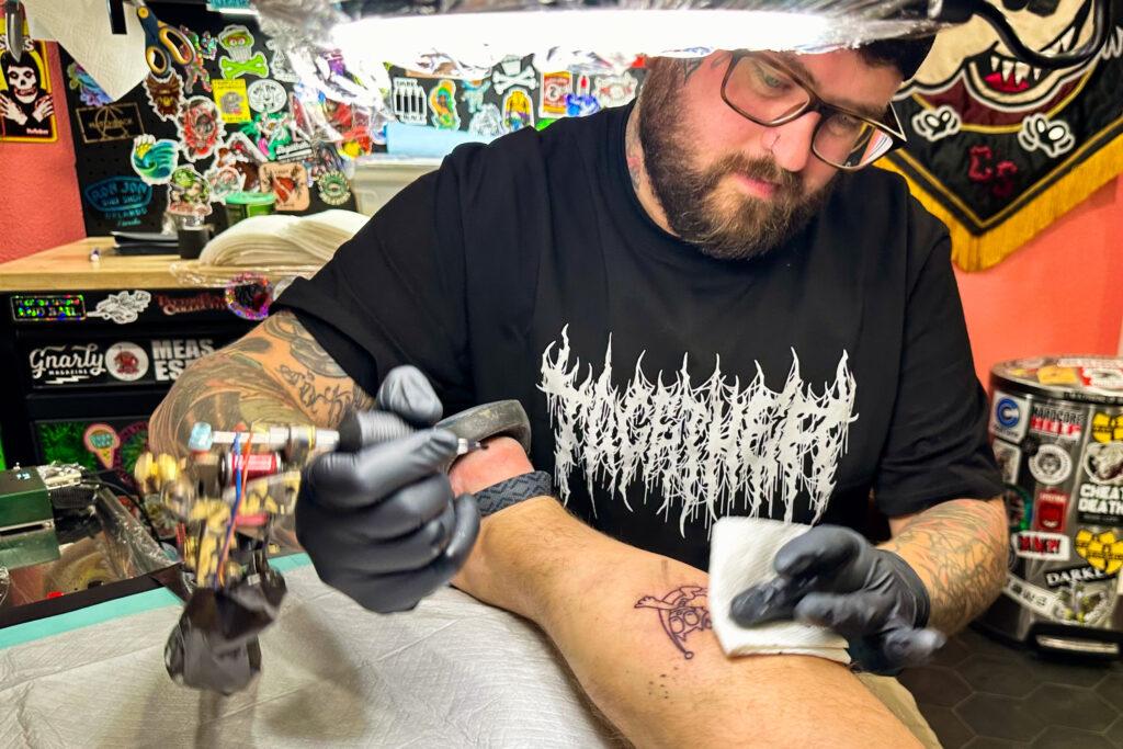 A bearded artist tattoos a cartoon cat on a customers calf