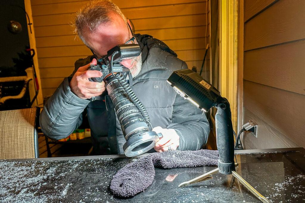 Man holding a camera with a telephoto lens takes pictures of microscopic snowflakes on a fluffy black sock as a background. The man is outside wearing a winter jacket.