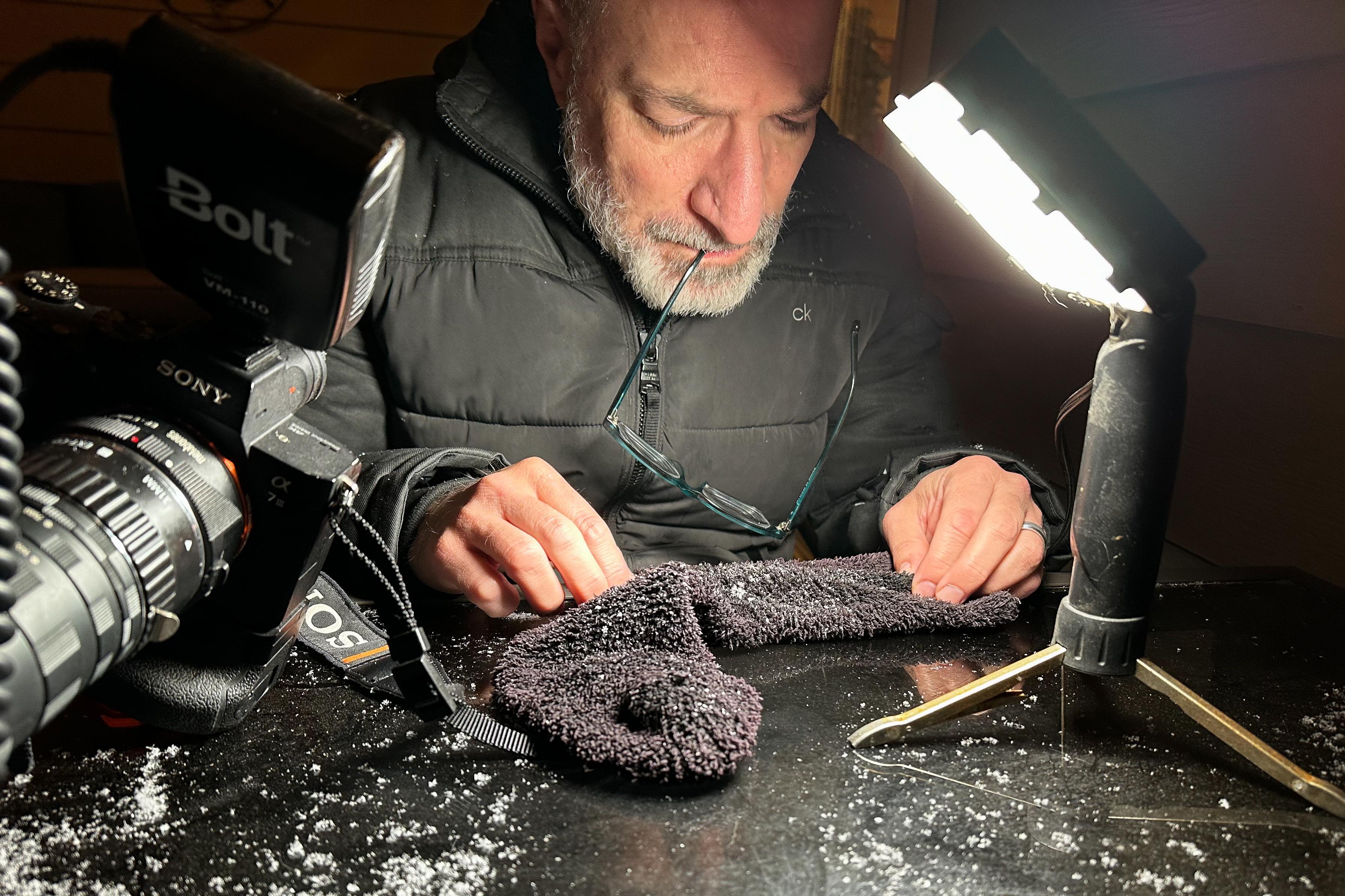 A man looks at a sock that has snowflakes on it under a photo light as he holds his glasses between his teeth. His camera sits next to him on a table.