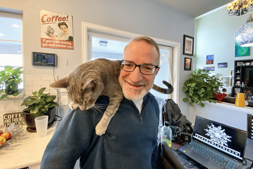 A man stands up at his computer desk smiling as a gray cat sits on his shoulder. The computer screen shows a picture of a snowflake.