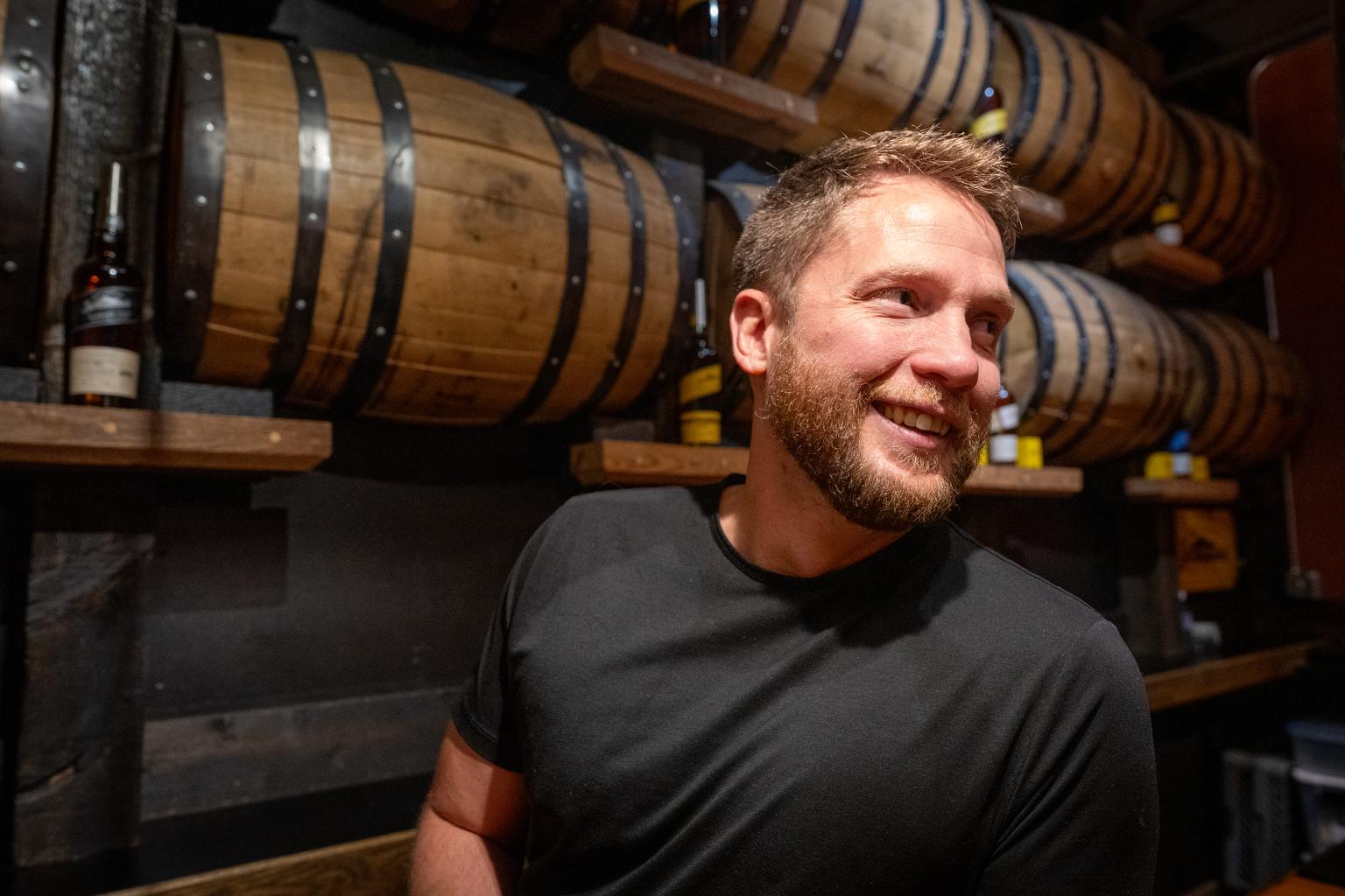 Justin Aden stands in front of wooden whiskey barrels.