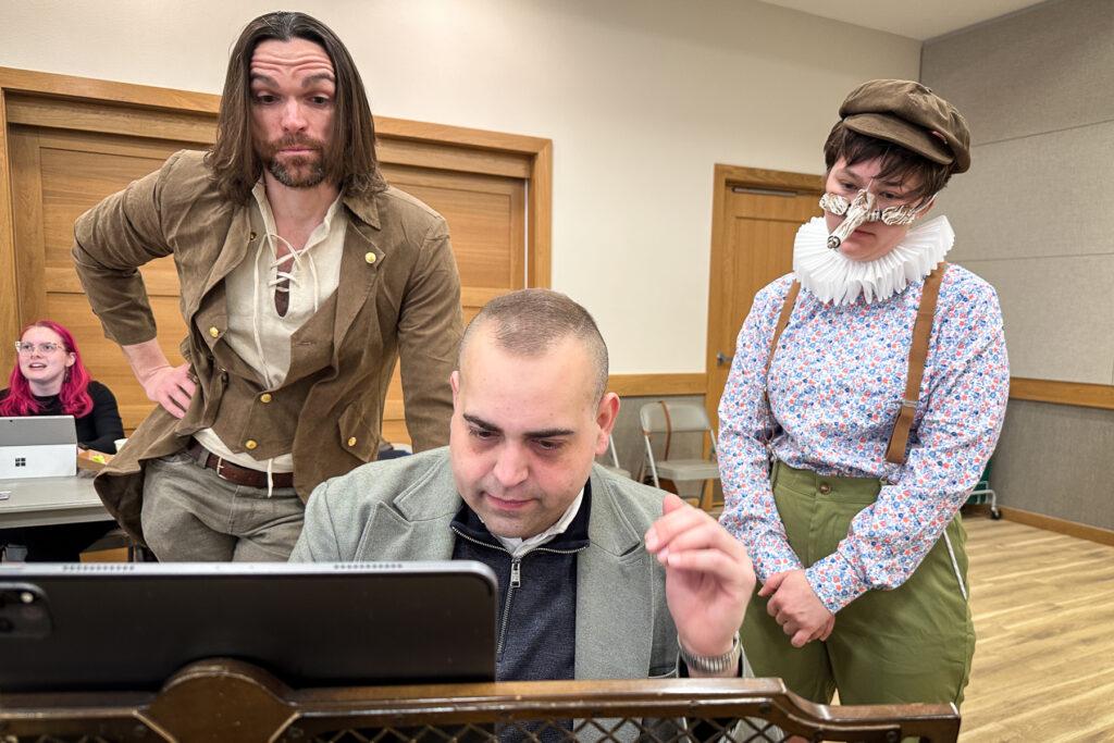 Two artists in costume, Tyler Padgett and Casey Klopp, stand behind music director Mario Barbosa who is seated at a piano.